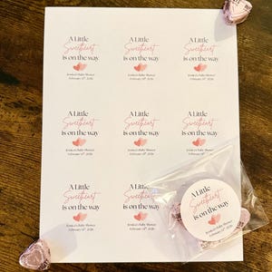 May include: A white sheet of paper printed with the text "A Little Sweetheart is on the way" in pink script, with pink heart graphics. A small bag of heart-shaped sweets and a few individual sweets are also visible.