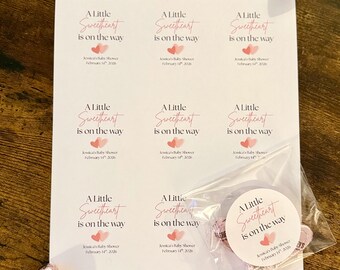 Può includere: Un foglio di carta bianco stampato con la scritta "A Little Sweetheart is on the way" in rosa, con grafiche di cuori rosa. È visibile anche un piccolo sacchetto di caramelle a forma di cuore e alcune caramelle singole.
