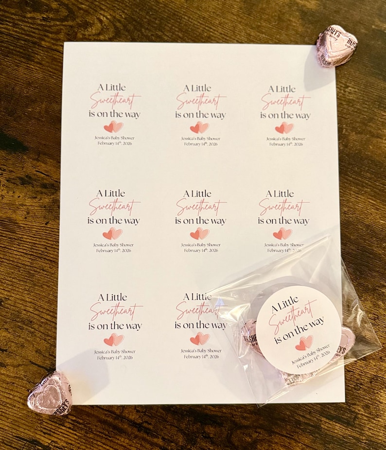 May include: A white sheet of paper printed with the text "A Little Sweetheart is on the way" in pink script, with pink heart graphics. A small bag of heart-shaped sweets and a few individual sweets are also visible.