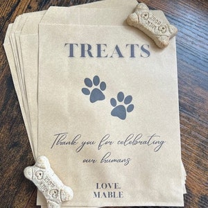 May include: Kraft paper treat bags with grey text reading "TREATS", paw prints, and "Thank you for celebrating our humans". The bags also have "LOVE. MABLE" printed on them. Two bone-shaped dog biscuits are placed on the bags.