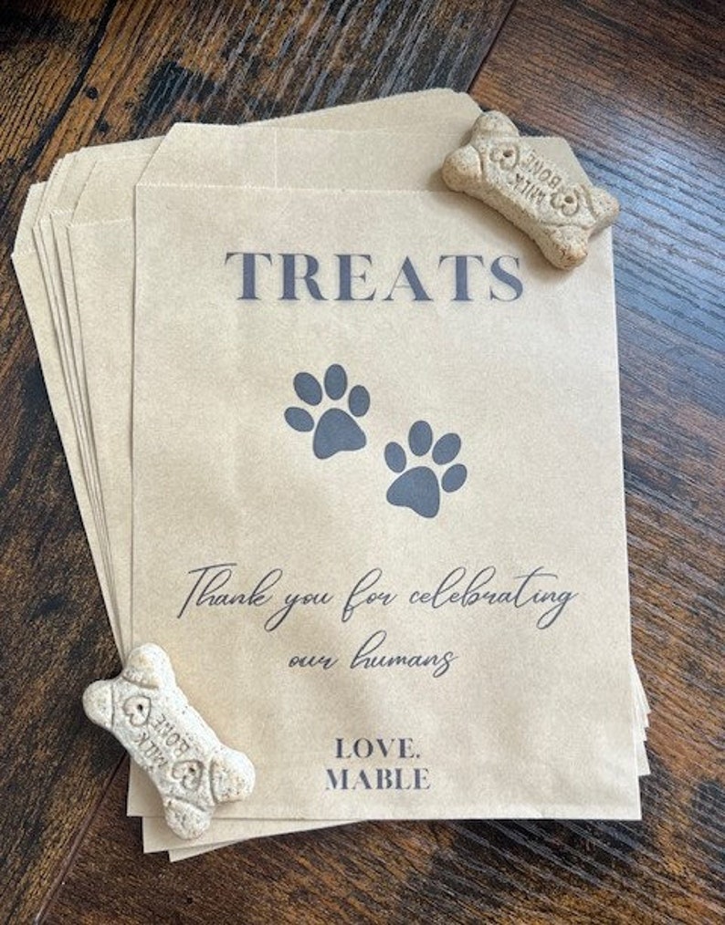 May include: Kraft paper treat bags with grey text reading "TREATS", paw prints, and "Thank you for celebrating our humans". The bags also have "LOVE. MABLE" printed on them. Two bone-shaped dog biscuits are placed on the bags.