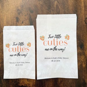 May include: Two white paper treat bags with the text "Two Little Cuties are on the way!" in orange and black. The bags also include the text "Brittany & Kyle's Baby Shower 08.30.2026".