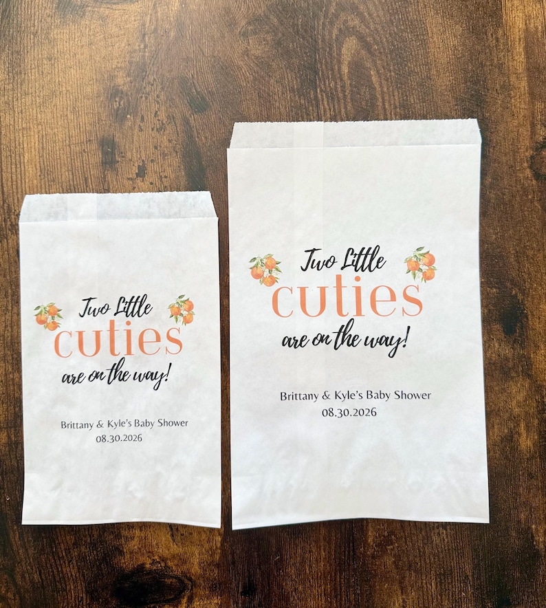 May include: Two white paper treat bags with the text "Two Little Cuties are on the way!" in orange and black. The bags also include the text "Brittany & Kyle's Baby Shower 08.30.2026".