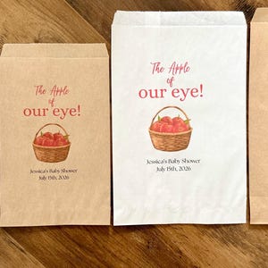 May include: Four rectangular paper favor bags in white and kraft brown colors. Each bag features a printed illustration of a woven basket filled with red apples, the text "The Apple of our eye!", "Jessica's Baby Shower", and "July 19th, 2026".