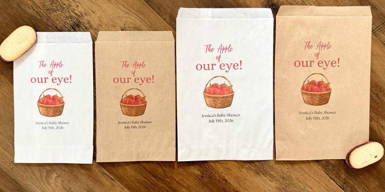 May include: Four rectangular paper favor bags in white and kraft brown colors. Each bag features a printed illustration of a woven basket filled with red apples, the text "The Apple of our eye!", "Jessica's Baby Shower", and "July 19th, 2026".