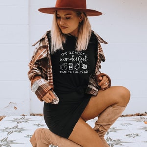 Puede incluir: Camiseta negra con el texto "It's the most wonderful time of the year" y gr&aacute;ficos tem&aacute;ticos de oto&ntilde;o. La persona lleva un sombrero marr&oacute;n y una camisa a cuadros. El atuendo se completa con un vestido negro y botas color canela.