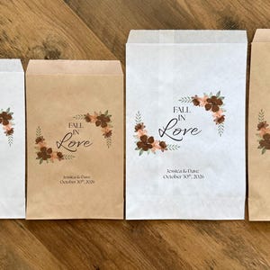 Puede incluir: Cuatro bolsas de papel rectangulares en blanco y marr&oacute;n kraft, con un dise&ntilde;o floral y el texto "FALL IN LOVE" y "Jessica & Dave October 30th, 2026". A la izquierda de las bolsas hay una galleta de chocolate con glaseado blanco.