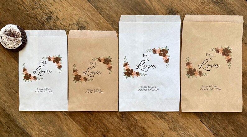 Puede incluir: Cuatro bolsas de papel rectangulares en blanco y marr&oacute;n kraft, con un dise&ntilde;o floral y el texto "FALL IN LOVE" y "Jessica & Dave October 30th, 2026". A la izquierda de las bolsas hay una galleta de chocolate con glaseado blanco.