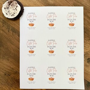 Op de afbeelding: Een wit vel papier met negen identieke rechthoekige stickers met de tekst A LITTLE Cutie Pie IS ON THE WAY! boven een kleine illustratie van een taartje en de tekst Jessica's Baby Shower August 29th, 2026 onder het taartje.