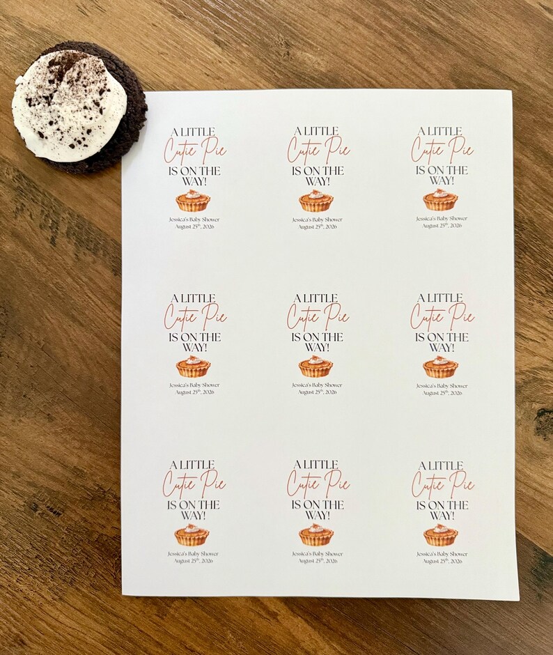 Op de afbeelding: Een wit vel papier met negen identieke rechthoekige stickers met de tekst A LITTLE Cutie Pie IS ON THE WAY! boven een kleine illustratie van een taartje en de tekst Jessica's Baby Shower August 29th, 2026 onder het taartje.