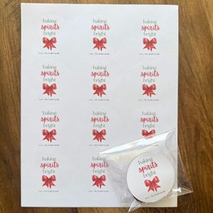Puede incluir: Una hoja de doce pegatinas circulares blancas con el texto rojo baking spirits bright sobre una ilustración de un lazo rojo y el texto negro Love, The Smith Family. Una pegatina adicional está en una bolsa de plástico.