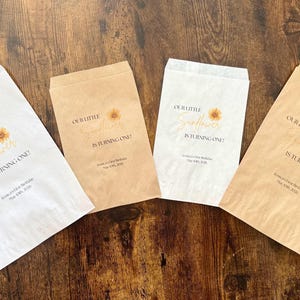 Puede incluir: Cuatro bolsas de papel para golosinas con el texto "OUR LITTLE Sunflower IS TURNING ONE!" en una fuente cursiva, con un gr&aacute;fico de girasol. Las bolsas son blancas y marrones, con el texto y el gr&aacute;fico en dorado y marr&oacute;n. Las bolsas est&aacute;n sobre una superficie de madera.