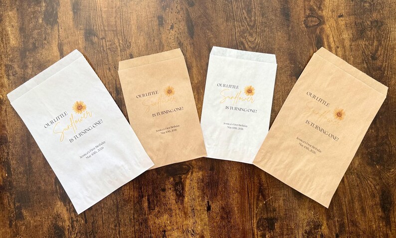 Puede incluir: Cuatro bolsas de papel para golosinas con el texto "OUR LITTLE Sunflower IS TURNING ONE!" en una fuente cursiva, con un gr&aacute;fico de girasol. Las bolsas son blancas y marrones, con el texto y el gr&aacute;fico en dorado y marr&oacute;n. Las bolsas est&aacute;n sobre una superficie de madera.