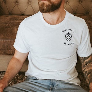 Può includere: T-shirt bianca con la scritta "DON'T WORRY BE HOPPY" e una grafica di luppolo. La maglietta è a girocollo. La maglietta è indossata da una persona.