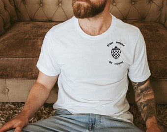Op de afbeelding: Wit T-shirt met de tekst "DON'T WORRY BE HOPPY" en een hop-afbeelding. Het T-shirt heeft een ronde hals. Het T-shirt wordt door een persoon gedragen.