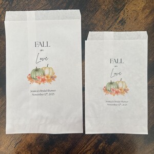May include: Two white paper treat bags with the text "FALL in Love" and a watercolour illustration of pumpkins and flowers. The text "Jessica's Bridal Shower November 12th, 2025" is printed below the illustration.