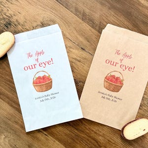 May include: Two rectangular paper favor bags, one white and one light brown, each printed with a red apple basket illustration and the text "The Apple of our eye! Jessica's Baby Shower July 15th, 2026". Two oval cookies are placed near the bags.