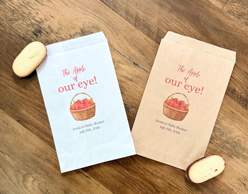 May include: Two rectangular paper favor bags, one white and one light brown, each printed with a red apple basket illustration and the text "The Apple of our eye! Jessica's Baby Shower July 15th, 2026". Two oval cookies are placed near the bags.