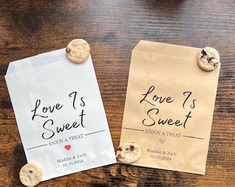 May include: Two paper treat bags, one white and one brown, with the text "Love Is Sweet" in black script. Each bag has a chocolate chip cookie attached. The white bag has a red heart and the names "Maddie & Zach" with a date.