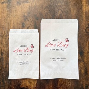 May include: Two white paper bags with the text "A Little Love Bug Is On The Way!" in pink script, with a butterfly graphic. The bags also include the text "Jessica's Baby Shower April 22nd, 2026."
