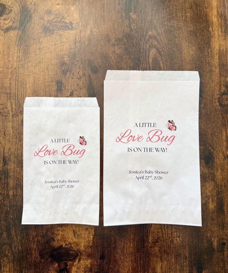 May include: Two white paper bags with the text "A Little Love Bug Is On The Way!" in pink script, with a butterfly graphic. The bags also include the text "Jessica's Baby Shower April 22nd, 2026."