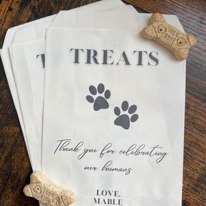 May include: White paper treat bags with the word "TREATS" printed in black, paw prints, and the text "Thank you for celebrating our humans. LOVE, MABLE." Two bone-shaped dog treats are also visible.