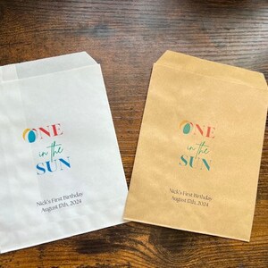 May include: Two paper treat bags, one white and one brown, with the text "ONE in the SUN" in colorful letters. Below the text is "Nick's First Birthday August 17th, 2024". The bags are for a birthday celebration.