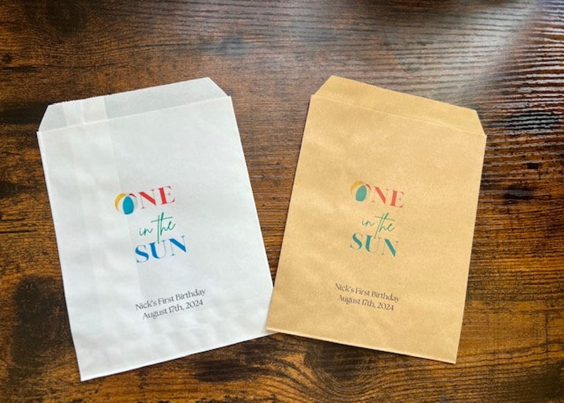 May include: Two paper treat bags, one white and one brown, with the text "ONE in the SUN" in colorful letters. Below the text is "Nick's First Birthday August 17th, 2024". The bags are for a birthday celebration.