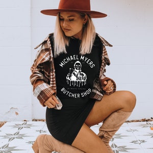 May include: Black t-shirt with the text "MICHAEL MYERS BUTCHER SHOP" in a white circular design. The shirt is worn with a brown plaid jacket, a black dress, and a brown hat. The image is taken outdoors.
