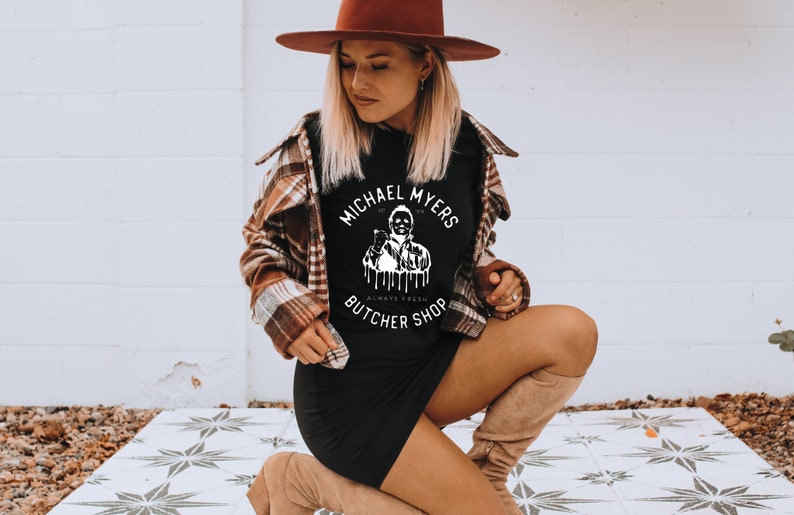May include: Black t-shirt with the text "MICHAEL MYERS BUTCHER SHOP" in a white circular design. The shirt is worn with a brown plaid jacket, a black dress, and a brown hat. The image is taken outdoors.