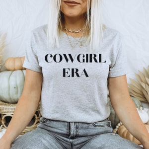 May include: A light gray t-shirt with the text "COWGIRL ERA" in a bold, black font. The shirt has short sleeves and a crew neck. The model is wearing blue jeans and gold necklaces.