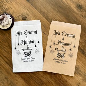 May include: Two rectangular paper favor bags, one white and one brown, each printed with black ink featuring the text We Created a Monster, a baby carriage illustration, spider webs, and spiders, plus Jessica's Baby Shower October 31, 2025.
