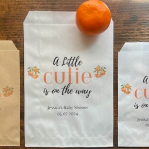 K&ouml;nnte beinhalten: Drei Papiert&uuml;ten mit dem Text "A Little Cutie is on the Way" und dem Datum 05.02.2024. Jede T&uuml;te hat eine kleine Orange oben drauf. Die T&uuml;ten sind in verschiedenen Gr&ouml;&szlig;en und Farben.