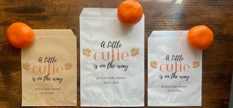 K&ouml;nnte beinhalten: Drei Papiert&uuml;ten mit dem Text "A Little Cutie is on the Way" und dem Datum 05.02.2024. Jede T&uuml;te hat eine kleine Orange oben drauf. Die T&uuml;ten sind in verschiedenen Gr&ouml;&szlig;en und Farben.