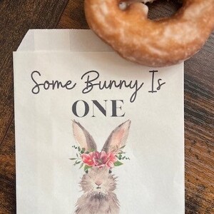 以下が含まれることがあります： 「Some Bunny Is ONE」の文字と、花の冠をつけたウサギの水彩イラストが描かれた白い紙袋。袋には「Violet's First Birthday March 25th 2024」の文字も含まれています。木製の表面には、グレーズされたドーナツが置かれています。