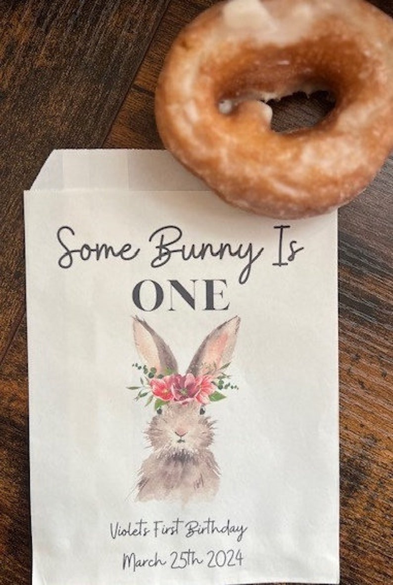 以下が含まれることがあります： 「Some Bunny Is ONE」の文字と、花の冠をつけたウサギの水彩イラストが描かれた白い紙袋。袋には「Violet's First Birthday March 25th 2024」の文字も含まれています。木製の表面には、グレーズされたドーナツが置かれています。