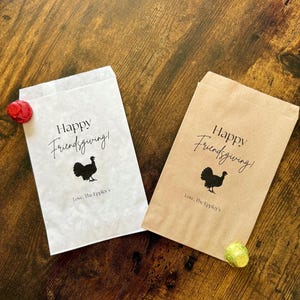 May include: Two paper treat bags, one white and one brown, with the text "Happy Friendsgiving!" and a turkey silhouette. The bags also include the text "Love, The Eppley's". A red and a green candy are next to the bags.