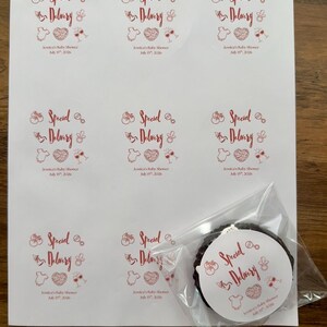 Op de afbeelding: Een vel met acht ronde witte stickers met rode tekst Special Delivery en Jessica's Baby Shower July 15th, 2026, met iconen van een rammelaar, fopspeen, rompertje, hart en wijnglazen. E&eacute;n sticker zit in een doorzichtig plastic zakje.