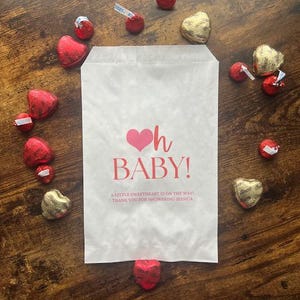 May include: A white paper favor bag featuring a pink heart icon and the red text Oh BABY! A LITTLE SWEETHEART IS ON THE WAY! THANK YOU FOR SHOWERING JESSICA. The bag is surrounded by red and gold heart-shaped chocolate candies on a wood surface.