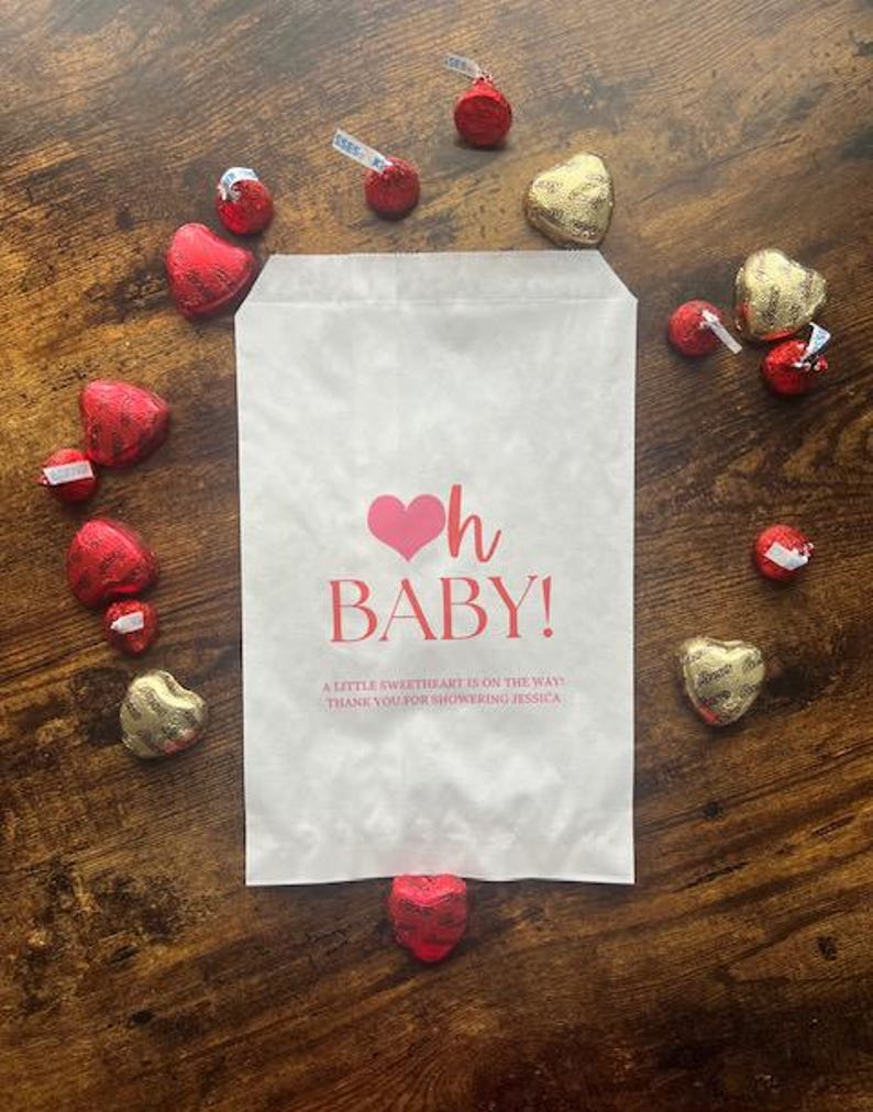 May include: A white paper favor bag featuring a pink heart icon and the red text Oh BABY! A LITTLE SWEETHEART IS ON THE WAY! THANK YOU FOR SHOWERING JESSICA. The bag is surrounded by red and gold heart-shaped chocolate candies on a wood surface.