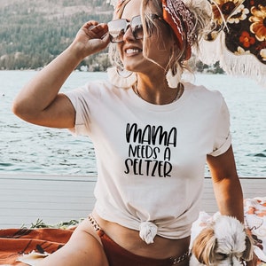 Puede incluir: Una camiseta blanca con el texto "Mama Needs a Seltzer" en escritura negra. La camiseta est&aacute; anudada a la cintura. La imagen est&aacute; tomada al aire libre con una masa de agua en el fondo.