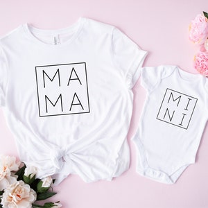 May include: White t-shirt with the word "MAMA" in a black square, next to a white baby onesie with "MINI" in a black square. The items are on a pink surface with pink and white flowers.