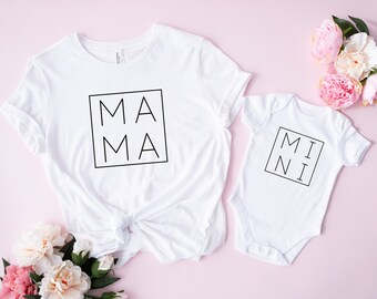May include: White t-shirt with the word "MAMA" in a black square, next to a white baby onesie with "MINI" in a black square. The items are on a pink surface with pink and white flowers.