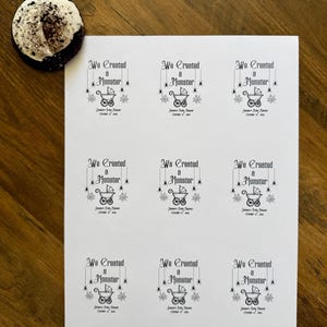 May include: A white sheet of paper featuring nine identical black printed labels. Each label displays the text We Created a Monster, a graphic of a baby carriage, and spider web accents, with the text Janna's Baby Shower October 31, 2024.