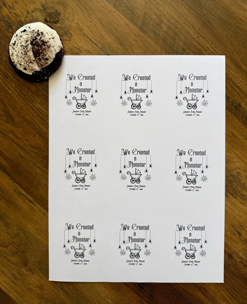 May include: A white sheet of paper featuring nine identical black printed labels. Each label displays the text We Created a Monster, a graphic of a baby carriage, and spider web accents, with the text Janna's Baby Shower October 31, 2024.