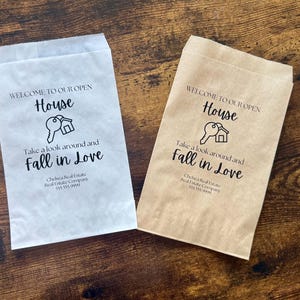 May include: Two paper bags, one white and one tan, with the text "WELCOME TO OUR OPEN HOUSE" and "Take a look around and Fall in Love" printed in black. A key and house graphic is also printed on the bags. The bags are for a real estate company.