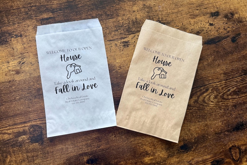 May include: Two paper bags, one white and one tan, with the text "WELCOME TO OUR OPEN HOUSE" and "Take a look around and Fall in Love" printed in black. A key and house graphic is also printed on the bags. The bags are for a real estate company.
