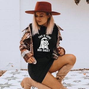 May include: Black t-shirt with the text "THIS IS SOME BOO SHEET" and a ghost wearing sunglasses. The person is wearing a brown hat and a plaid shirt. The person is also wearing a black dress and tan boots.