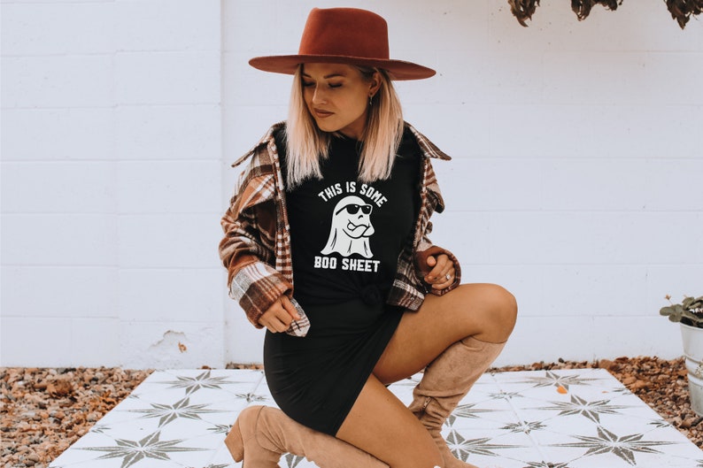 May include: Black t-shirt with the text "THIS IS SOME BOO SHEET" and a ghost wearing sunglasses. The person is wearing a brown hat and a plaid shirt. The person is also wearing a black dress and tan boots.
