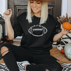 Puede incluir: Sudadera negra con el texto "gameday" sobre un gr&aacute;fico de f&uacute;tbol americano blanco. La imagen presenta calabazas y decoraci&oacute;n oto&ntilde;al, lo que sugiere un producto tem&aacute;tico de temporada o deportivo.
